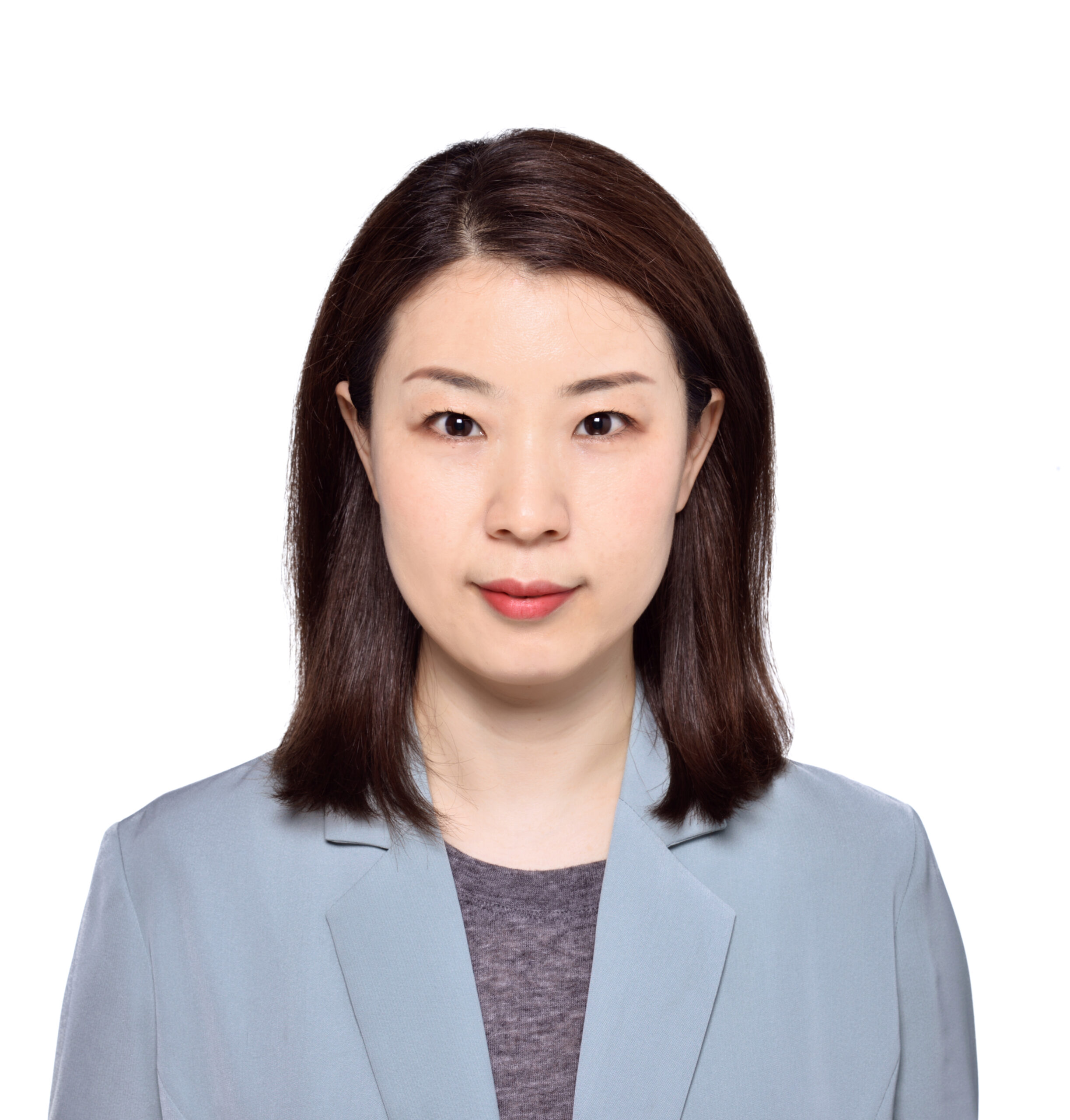 A woman with straight, shoulder-length dark hair wearing a light blue blazer over a gray top, facing forward with a neutral expression against a plain white background.